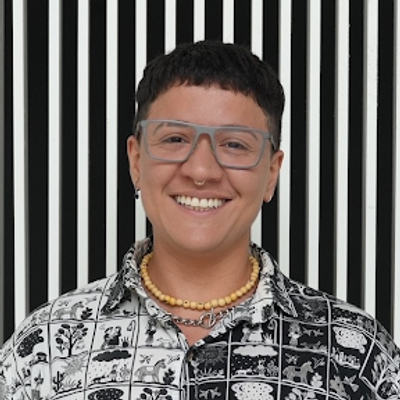 Smiling person with short dark hair wearing clear glasses, a patterned black-and-white shirt, and a beaded necklace, standing in front of a background with vertical black and white stripes.